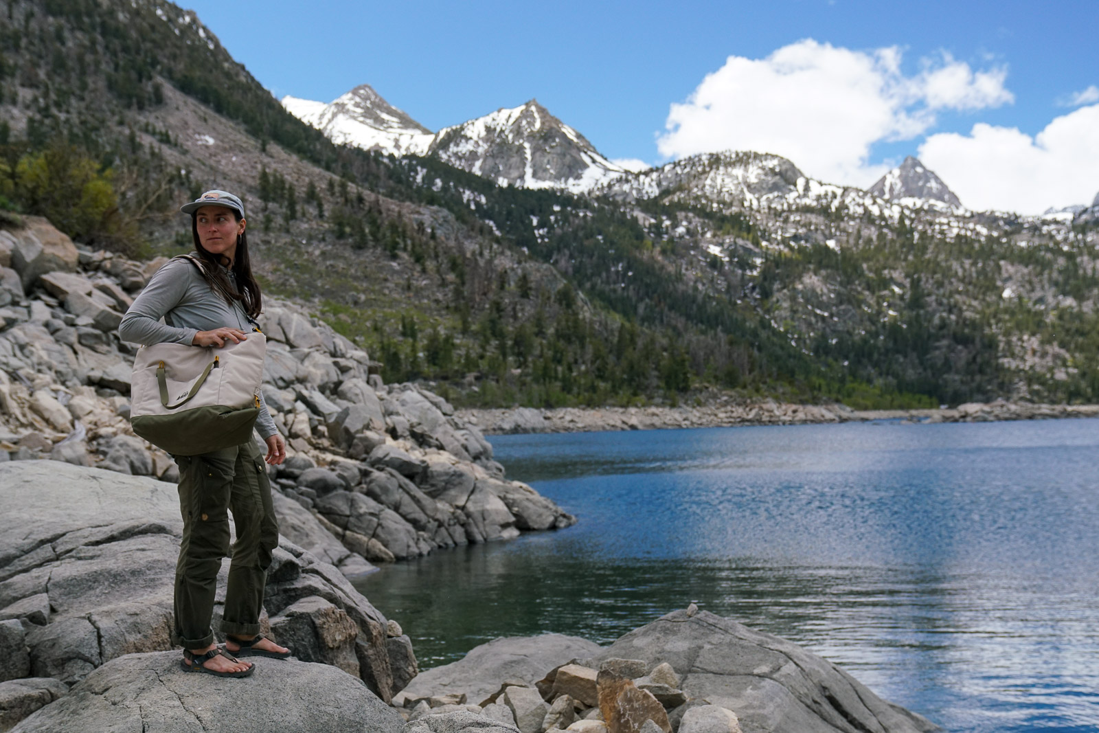 A person stands beside a lake bordered by mountains with a soft-sided cooler slung over their shoulder.