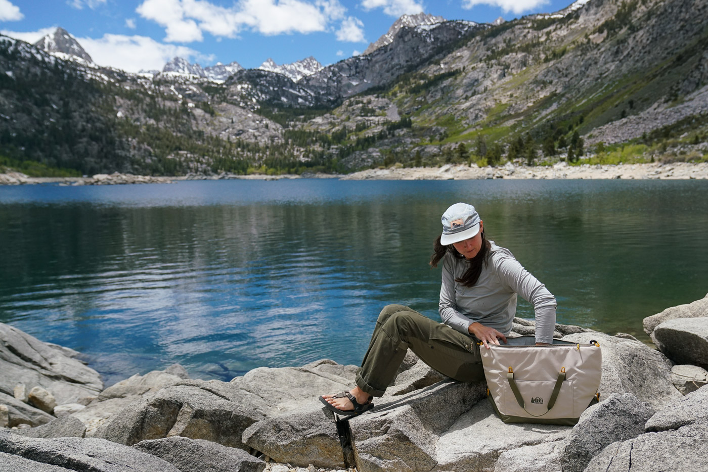 A person reaches into a soft-sided cooler while sitting on a rocky shoreline beside a mountain lake.