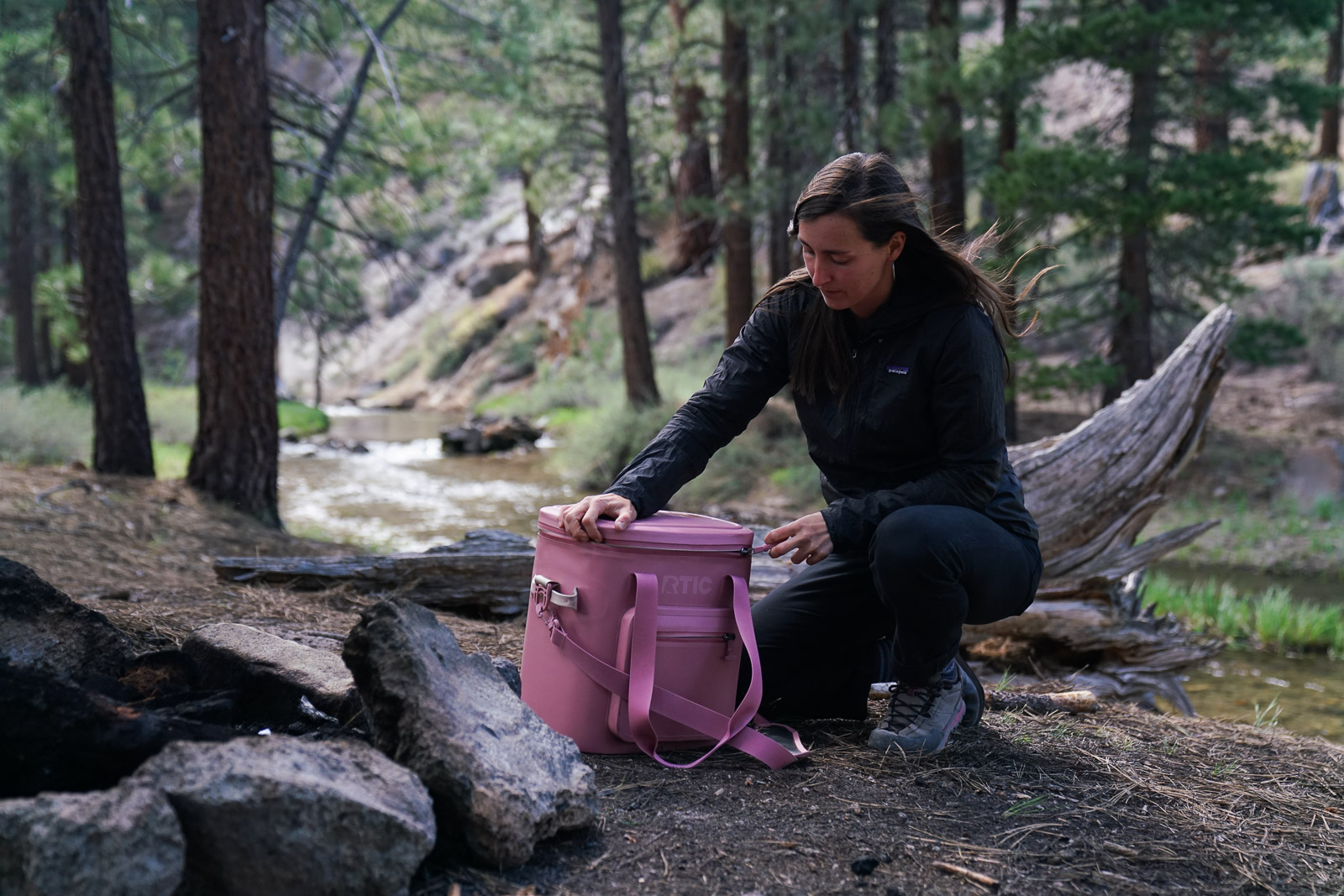 A person kneels beside a soft-sided cooler and unzips the lid with a forest and creek in the background.