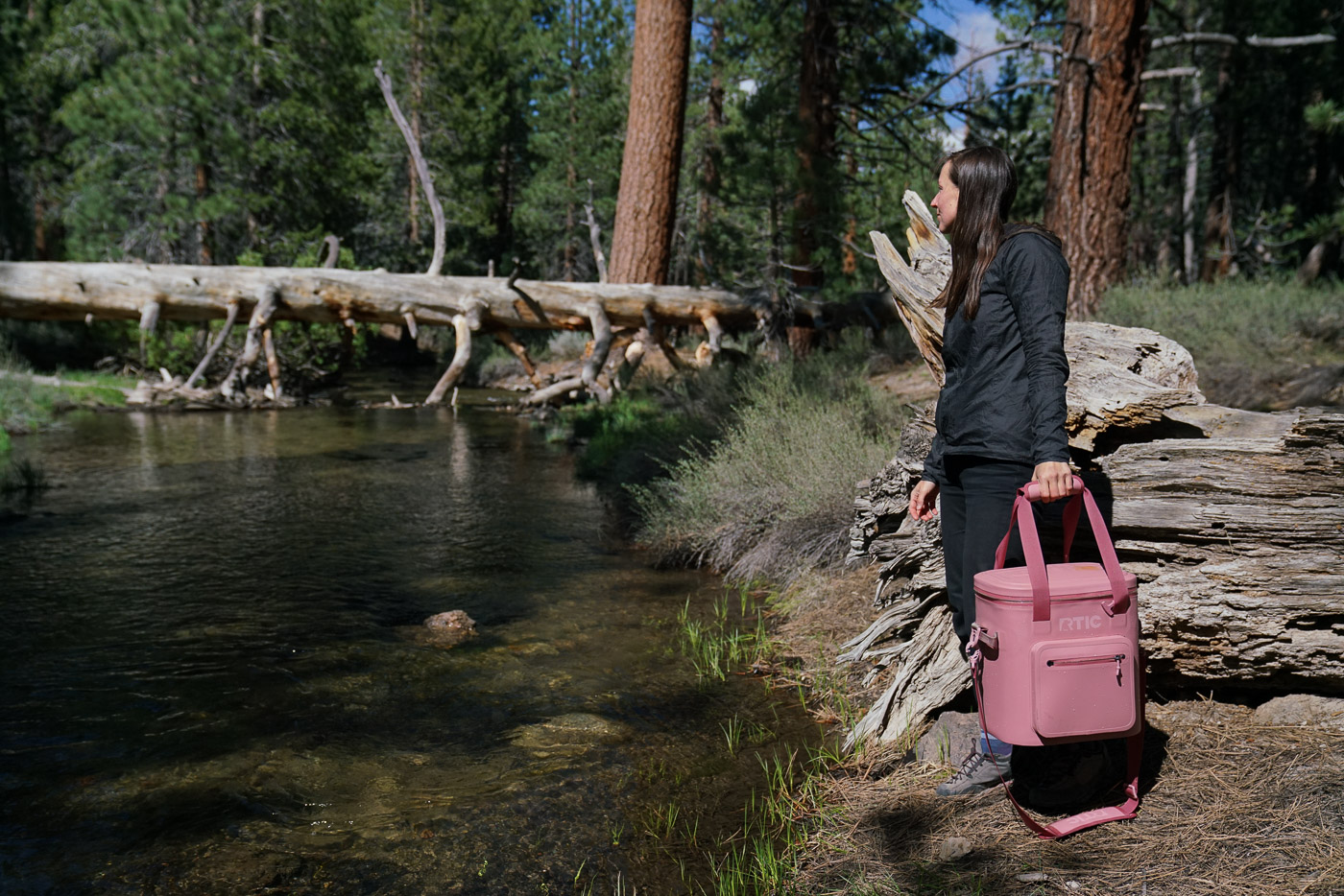 A person holds a soft-sided cooler while standing next to a creek.