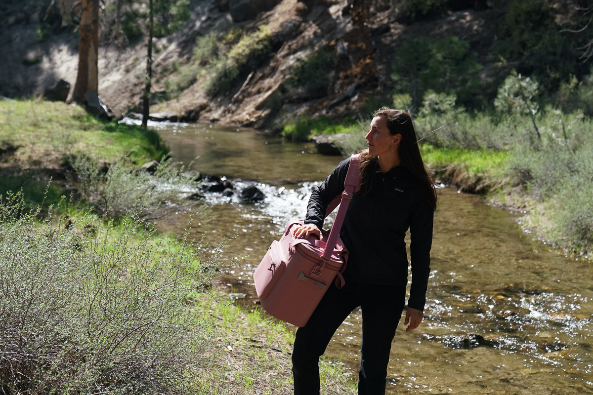 A person stands next to a creek with a soft-sided cooler slung over their shoulder.
