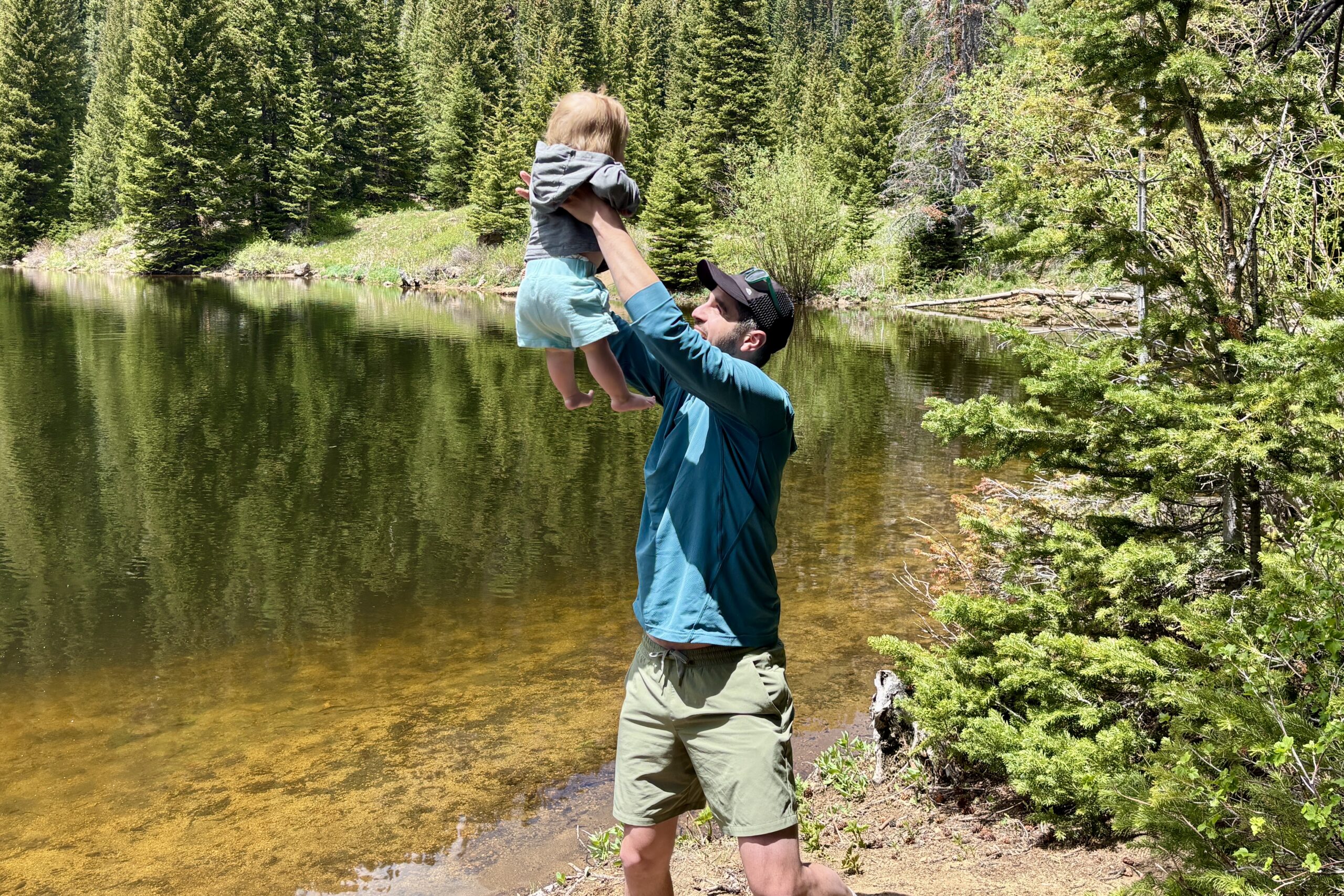 Father lifts infant into air standing on the edge of an alpine lake in a sunny spring day