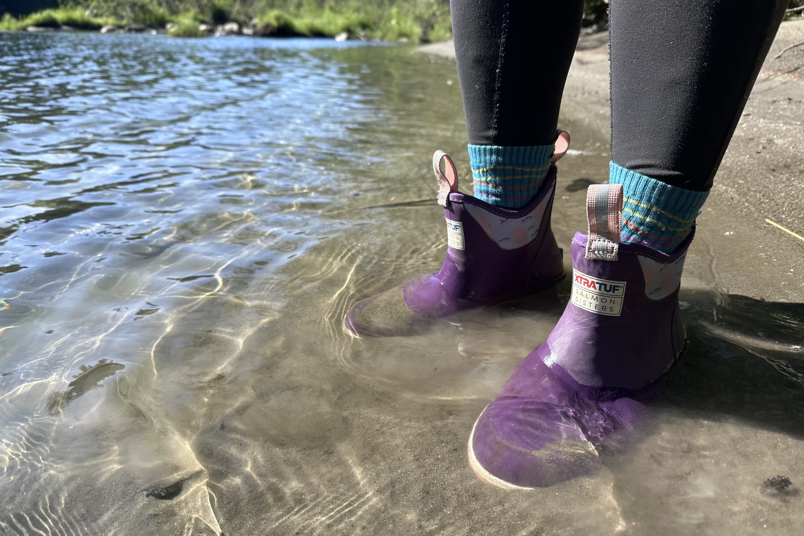 Close up of purple ankle rain boots standing just under ankle deep in a river next to a sandy beach.