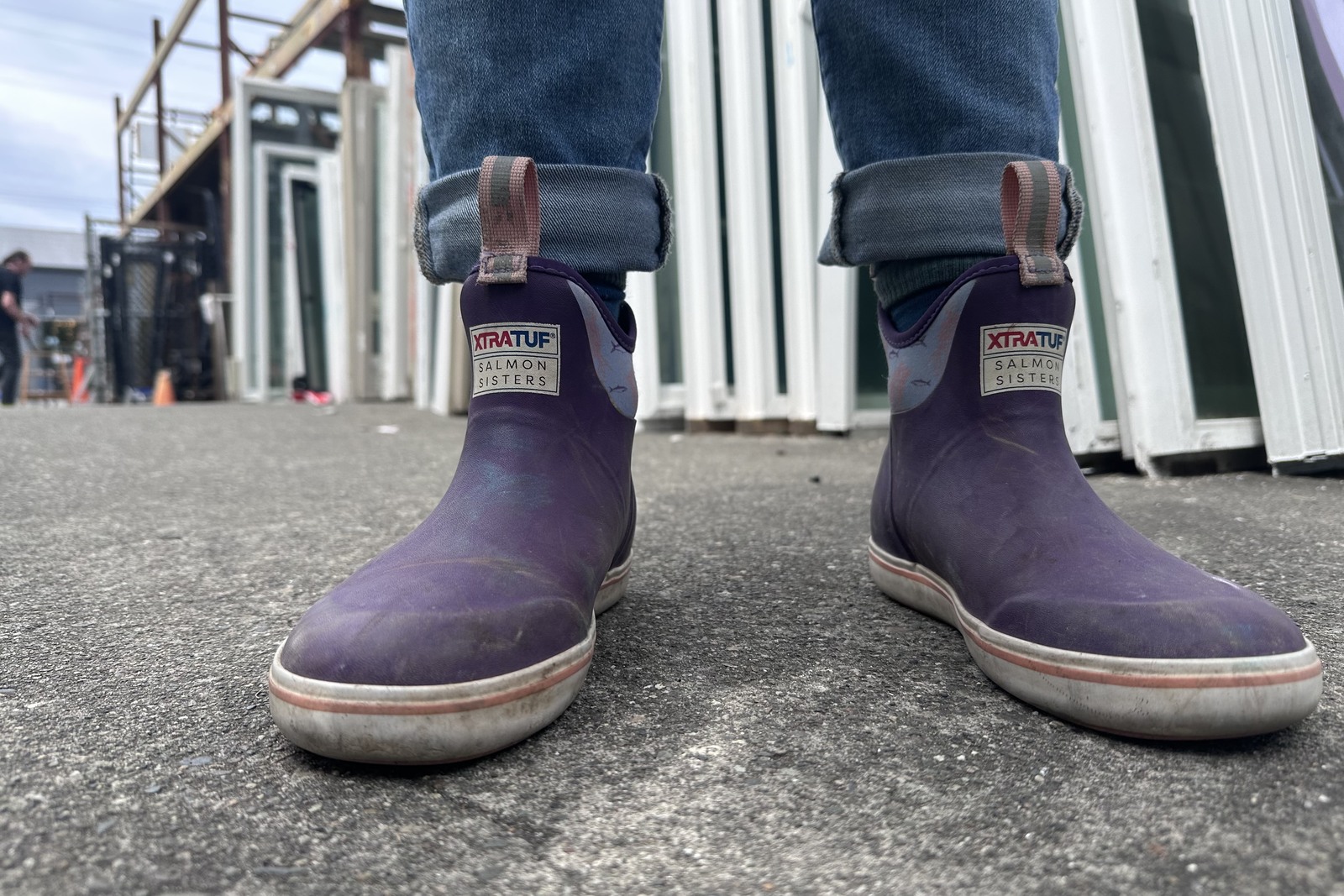 Close up of a pair of purple ankle boots standing on pavement next to a row of used windows.