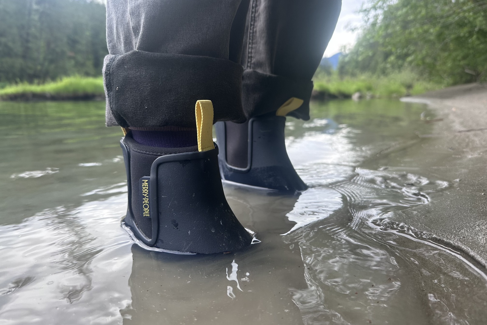 Close up of navy ankle boots standing in water near a small beach.