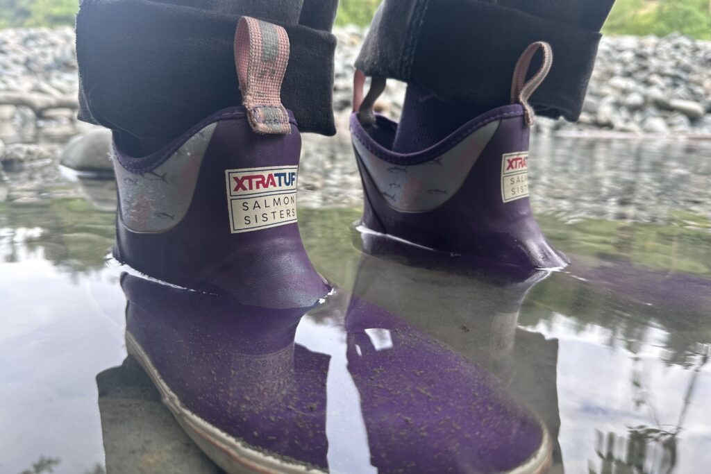 Close-up of purple ankle boots standing just under ankle deep in a shallow, rocky pool.