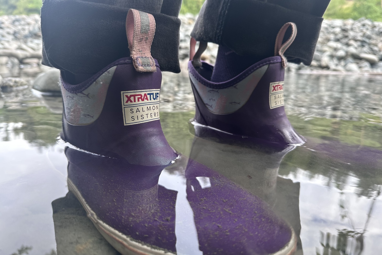 Close-up of purple ankle boots standing just under ankle deep in a shallow, rocky pool.