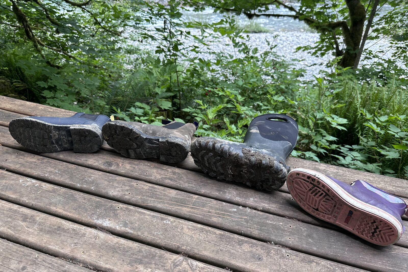 Four pairs of waterproof boots are lying on a wooden porch so that the bottom of each boot is visible to compare the treads.