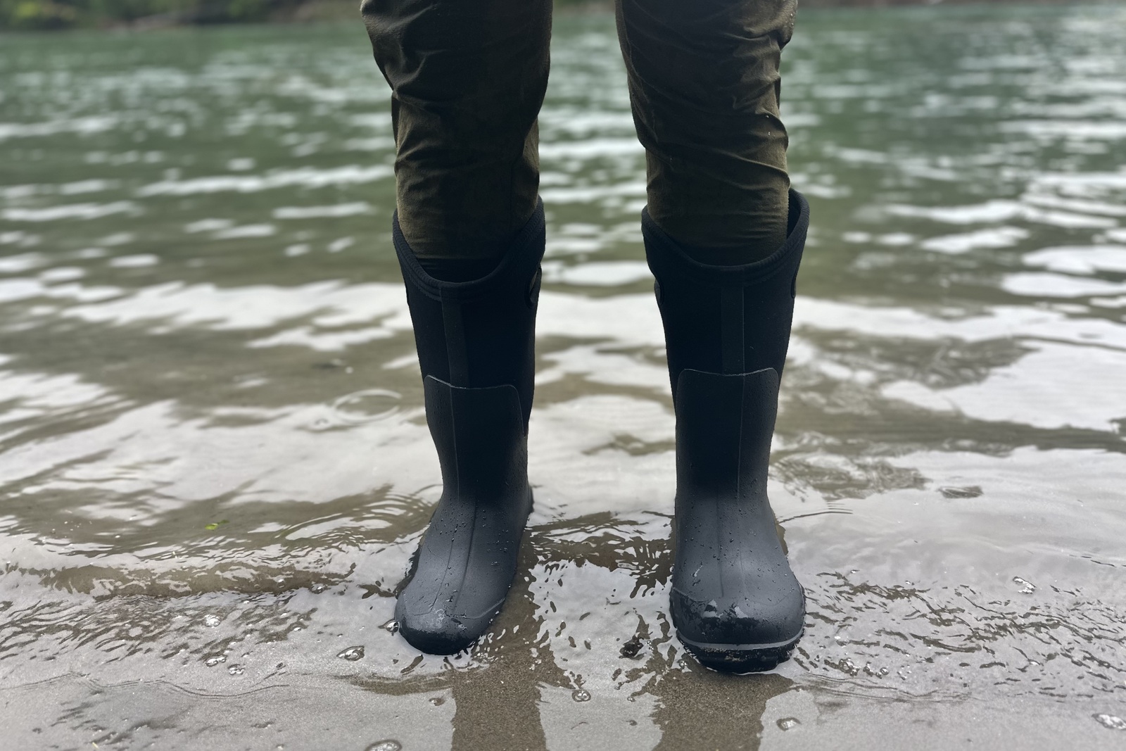 Close up of tall, black rubber boots with neoprene liners standing in shallow water on a beach.