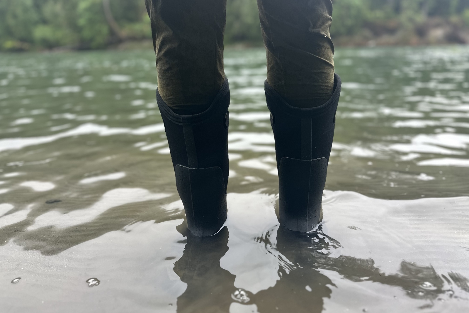Close up of tall black rubber rain boots with neoprene liners standing ankle deep in a sandy river.