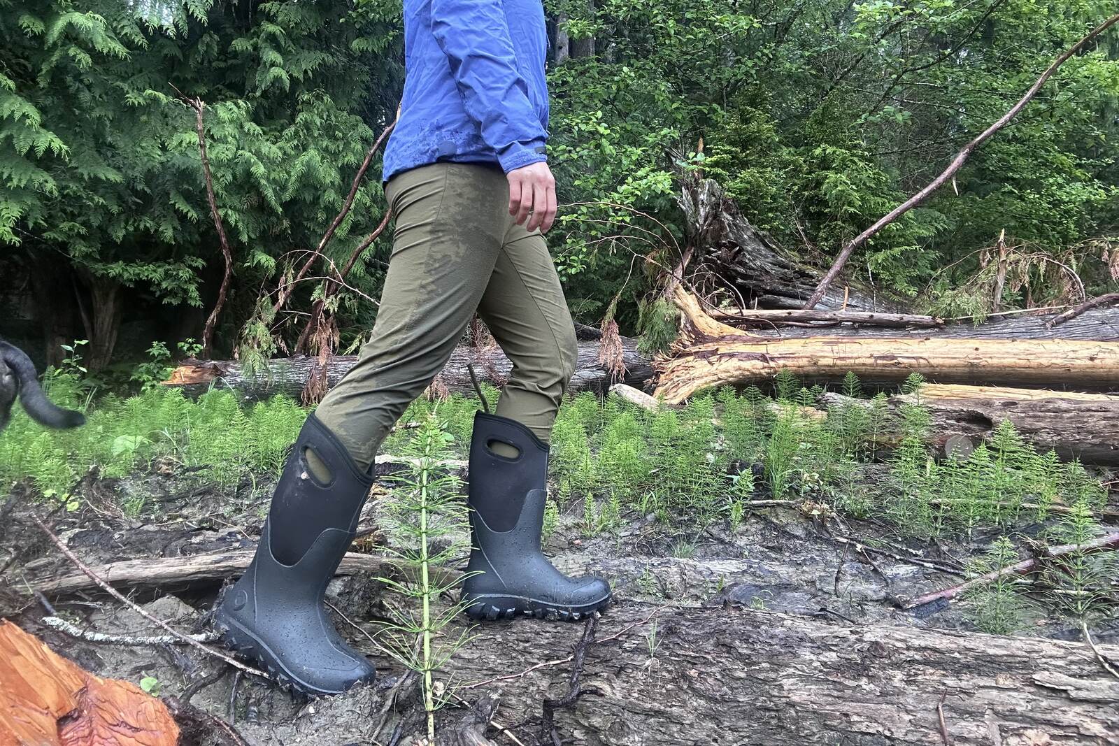 Photo of a person's lower half wearing tall black rain boots and walking through mud next to a forest.