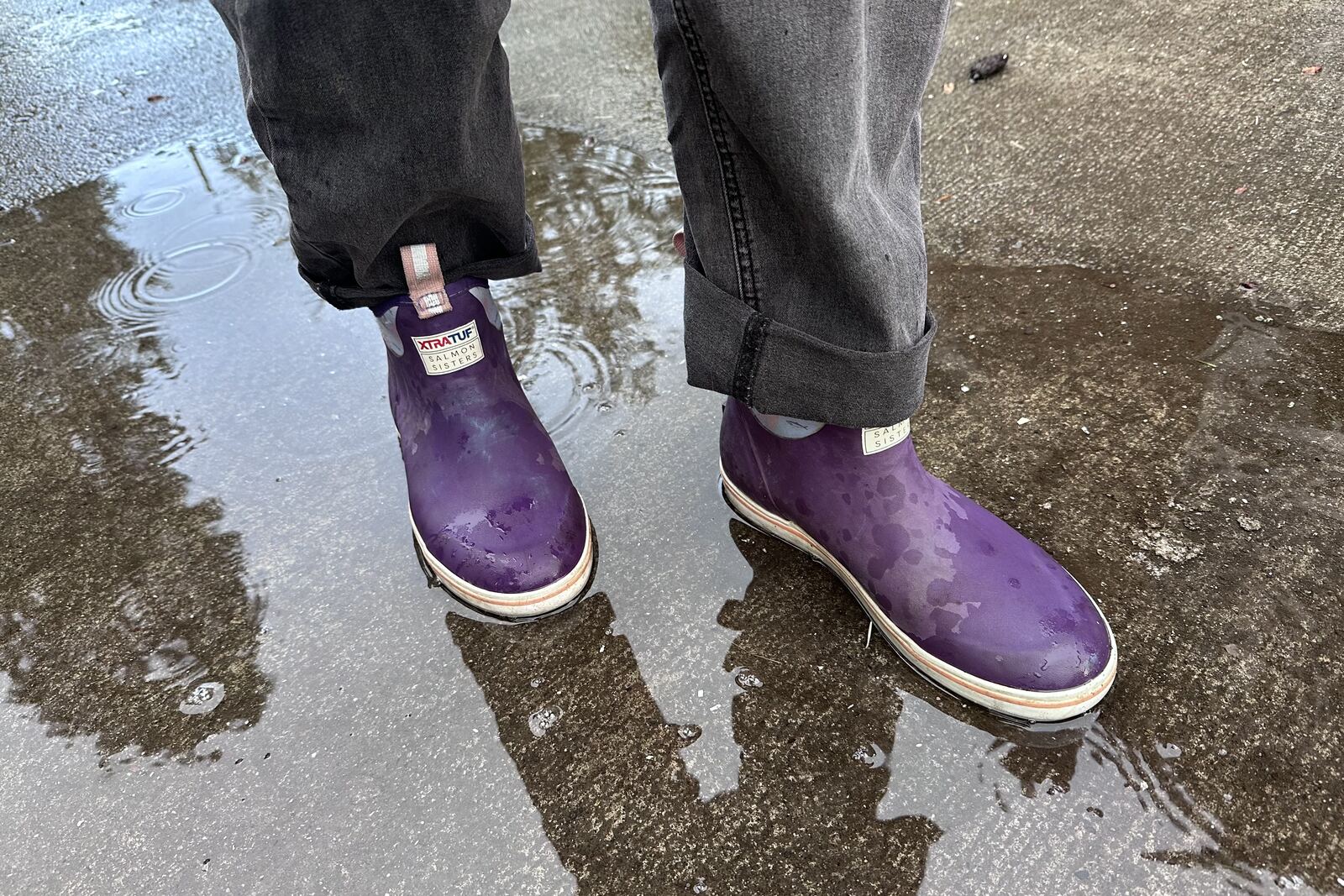 Close up of purple ankle rain boots standing in a puddle on pavement.