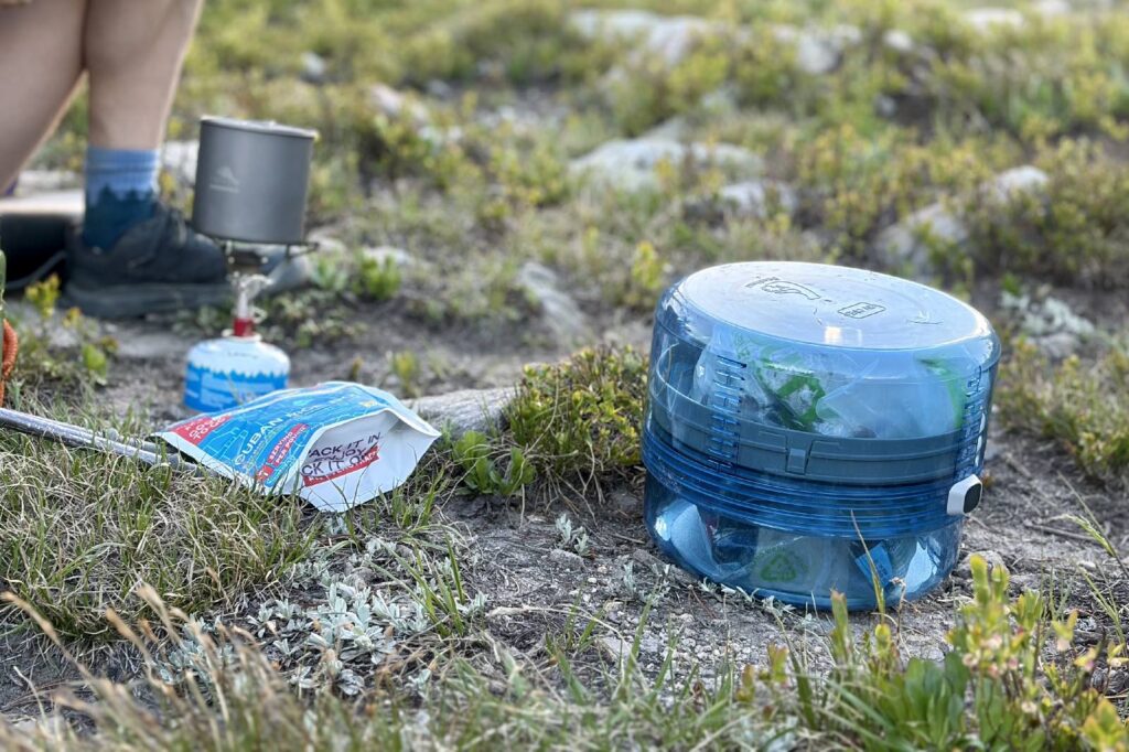 A clear blue bear canister sitting next to some backpacking cooking gear
