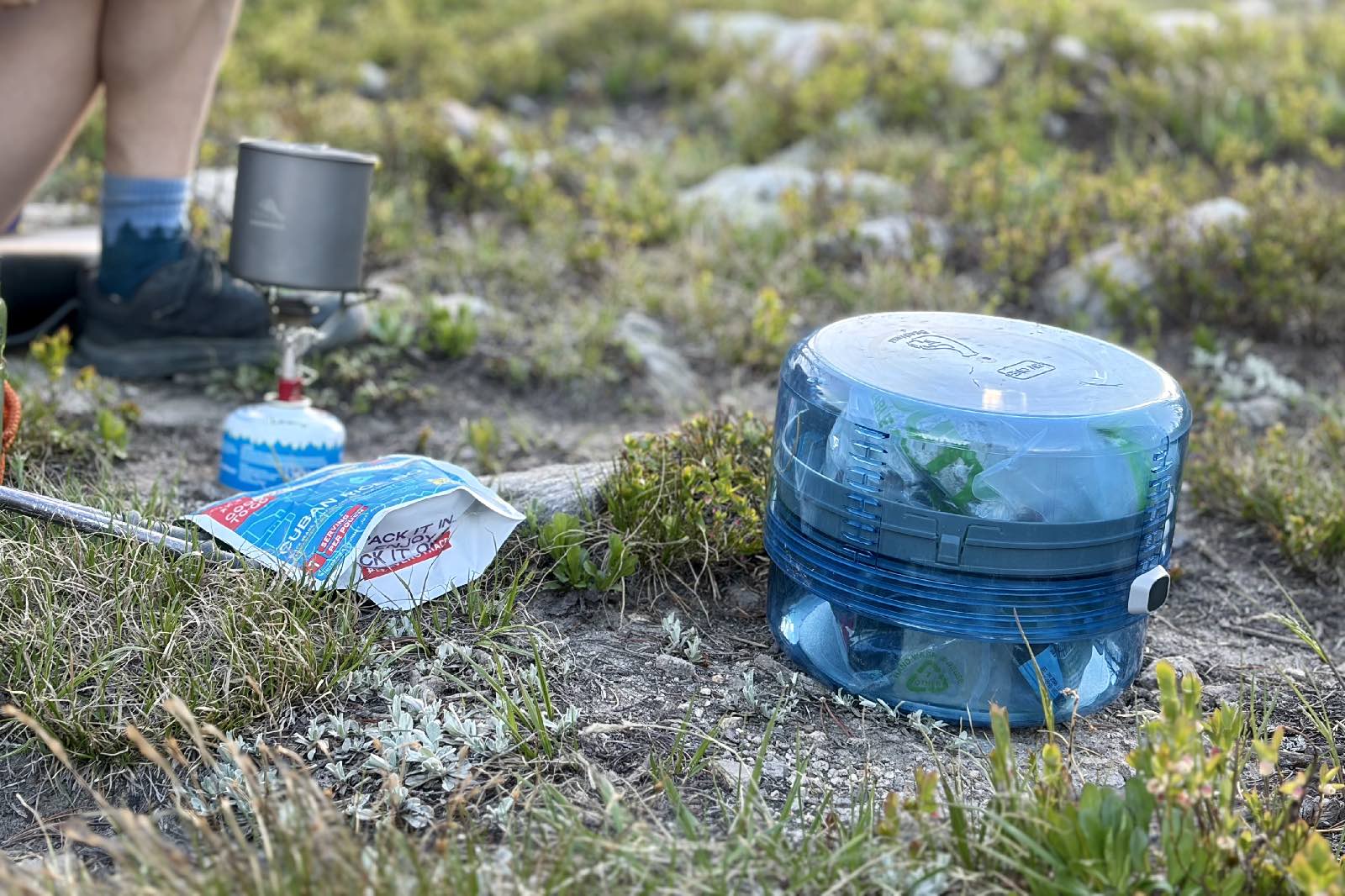 A clear blue bear canister sitting next to some backpacking cooking gear