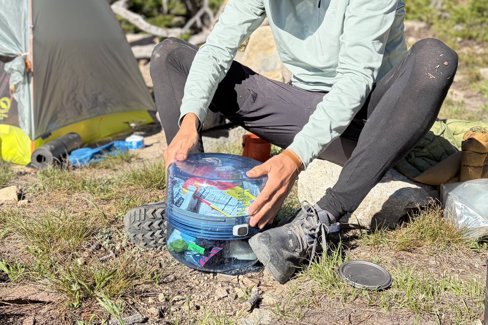 A hiker holds a bear canister with his feet while twisting apart the halves.