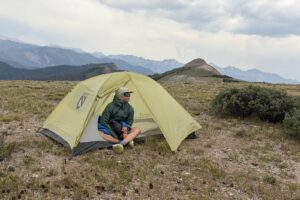 a backpacking tent pitched with a mountain background. a hiker sits inside.