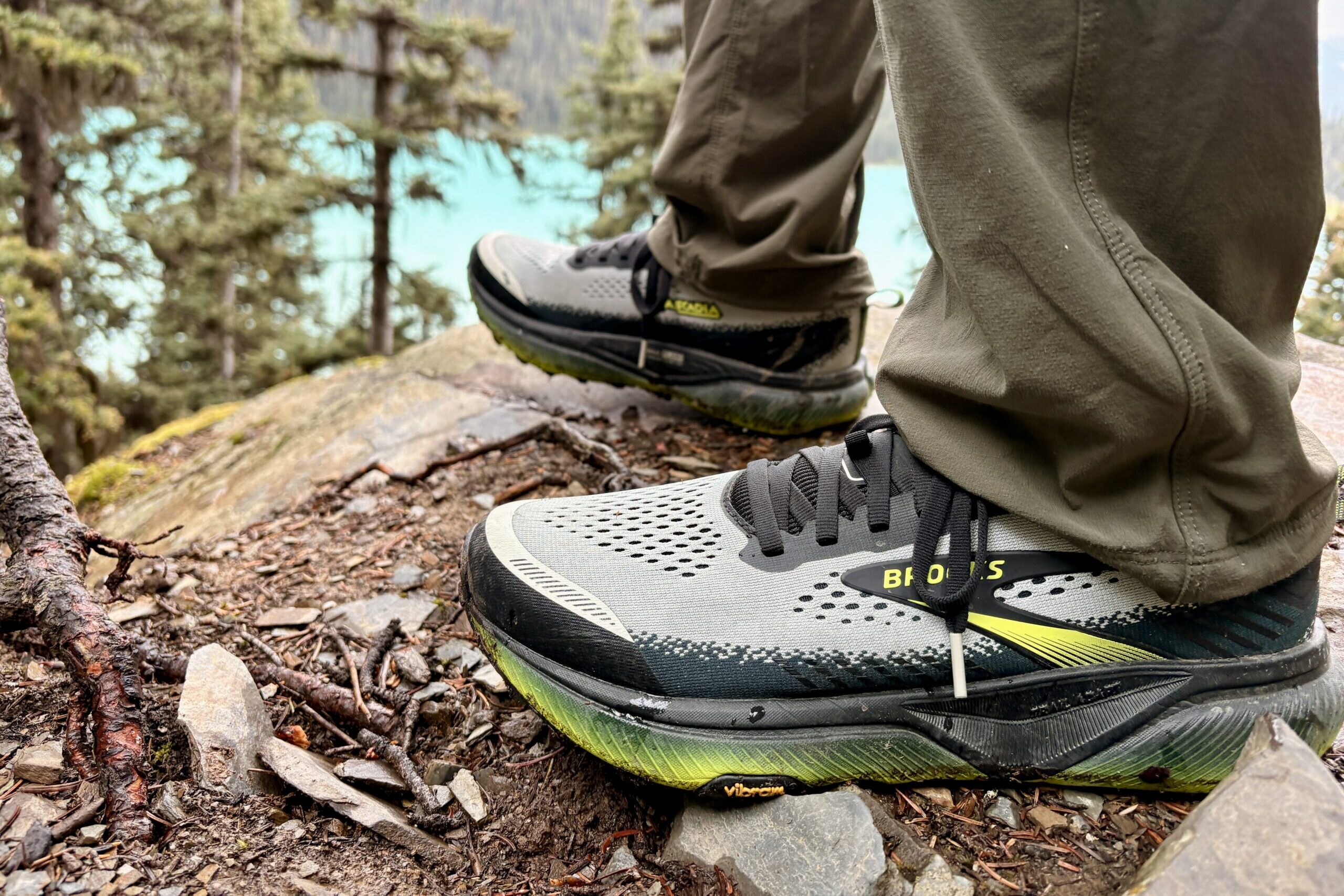 A closeup of a man's trail running shoe on dirt and rocks with a lake and forest in the background