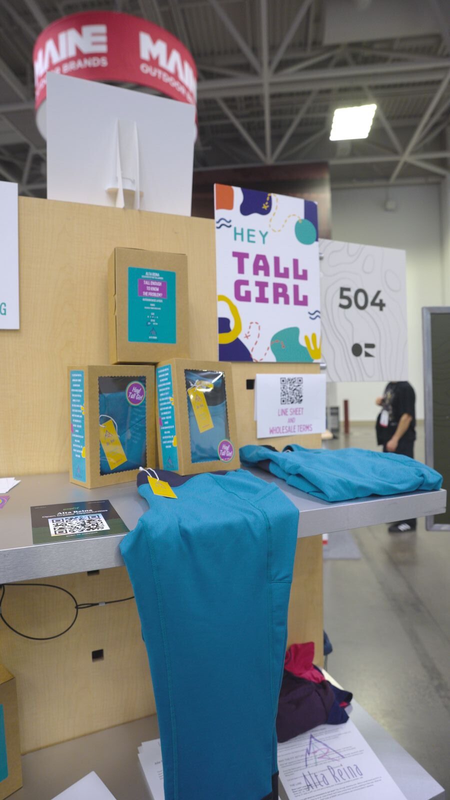 A set of teal base layers sit on a display that says "Hey Tall Girl" at a trade show.